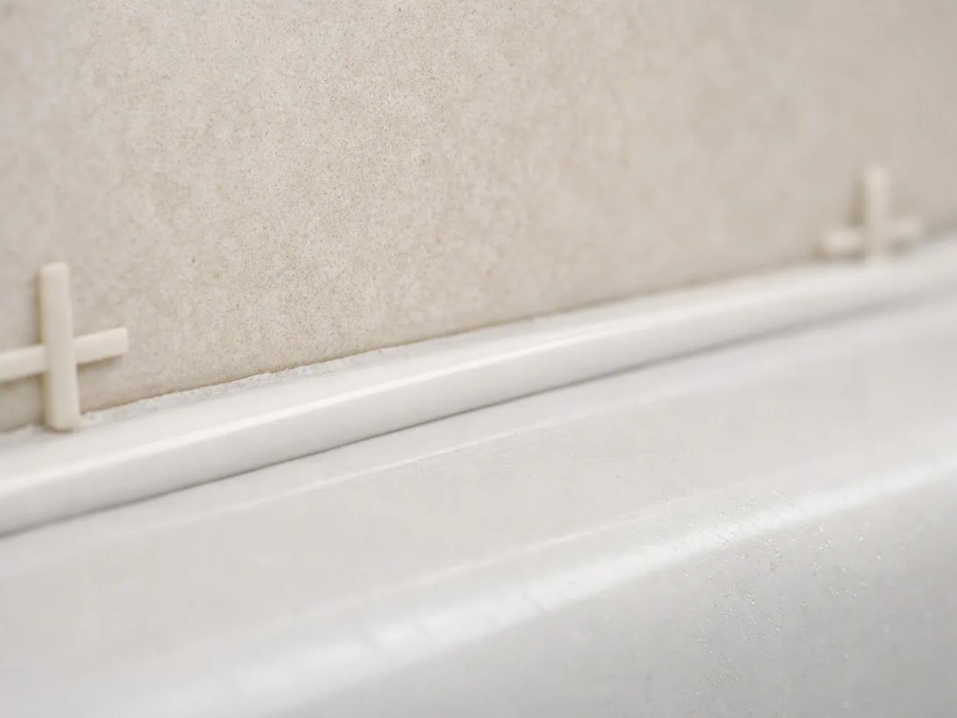 Close-up of a white bathtub edge beside a beige tiled wall with small spacer clips
