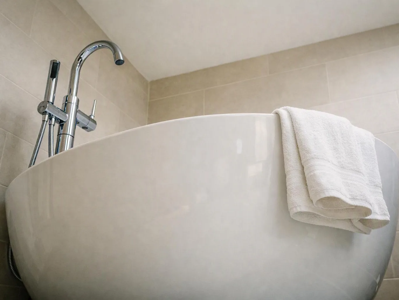 A white freestanding bathtub sits in a tiled bathroom, with a chrome faucet and a folded towel draped over the rim